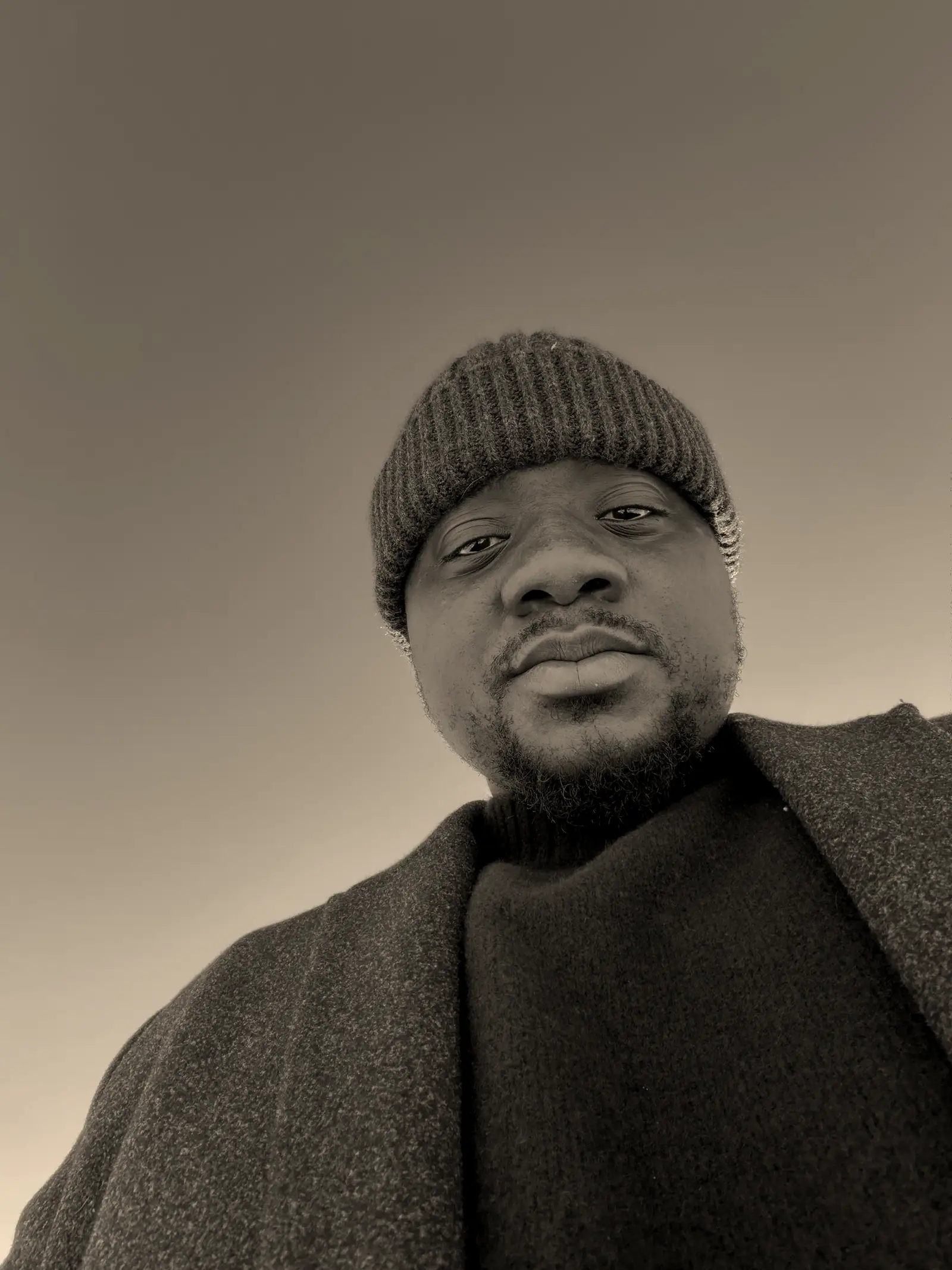David in a wool beanie and dark coat, looking down into the camera under a clear sky.